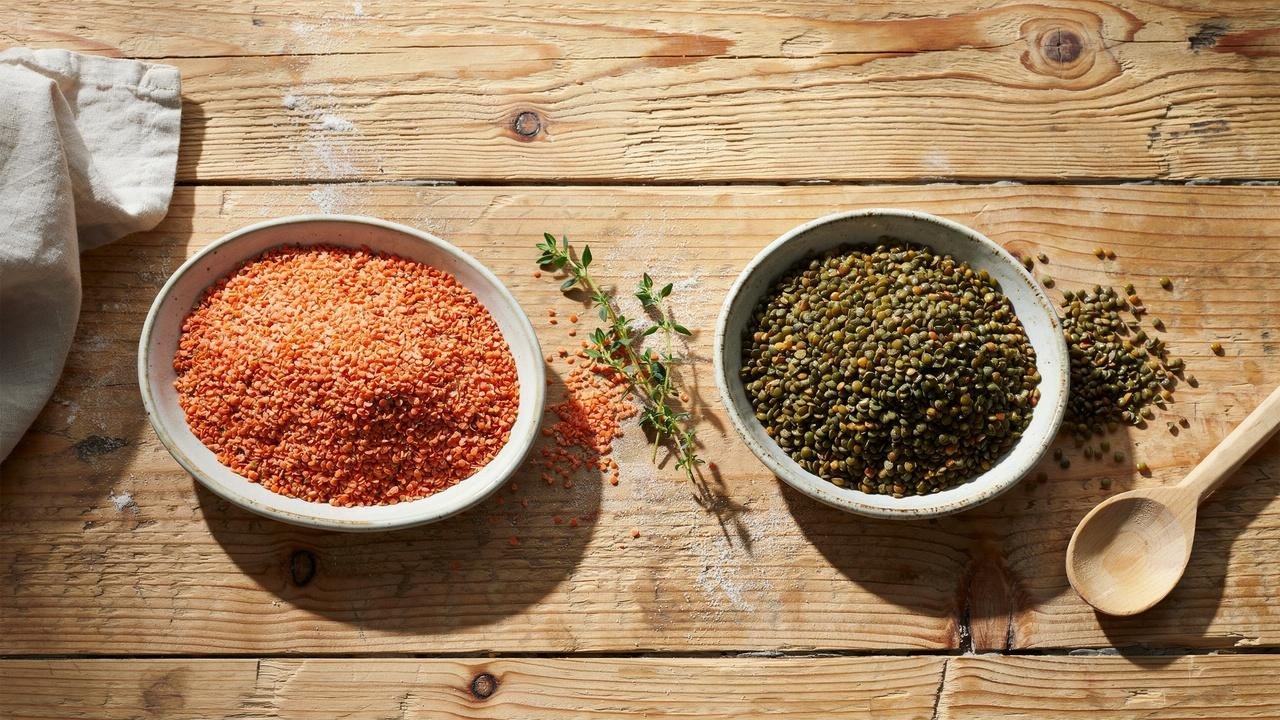 Two bowls on a rustic wooden table, one filled with red lentils and the other with green lentils.
