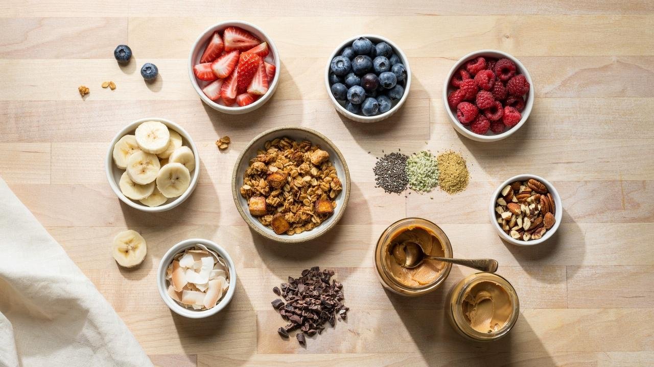 Small bowls of berries, granola, seeds, coconut flakes, cacao nibs, peanut butter, and nuts on a counter.