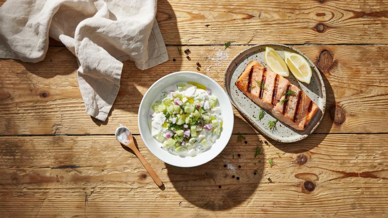 Creamy cucumber dill yogurt salad in a white ceramic bowl beside a plate of grilled baked salmon fillet.