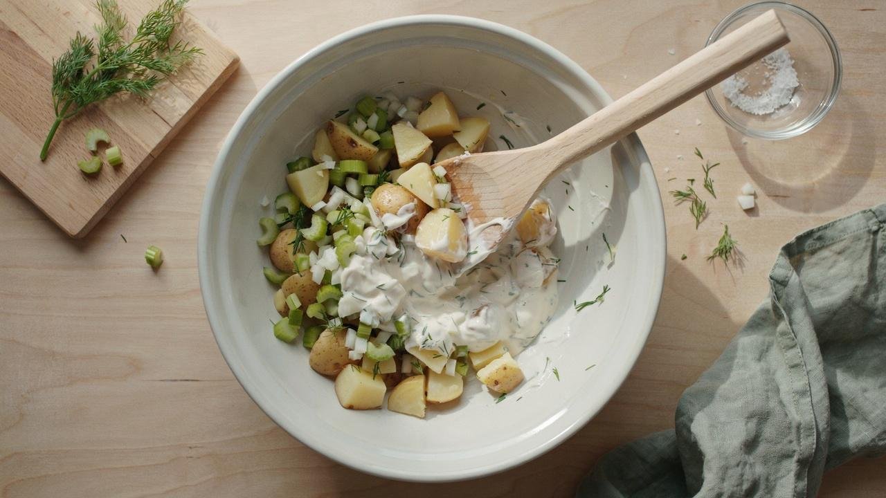 Cooked potatoes and chopped vegetables being gently mixed into Greek yogurt dressing.