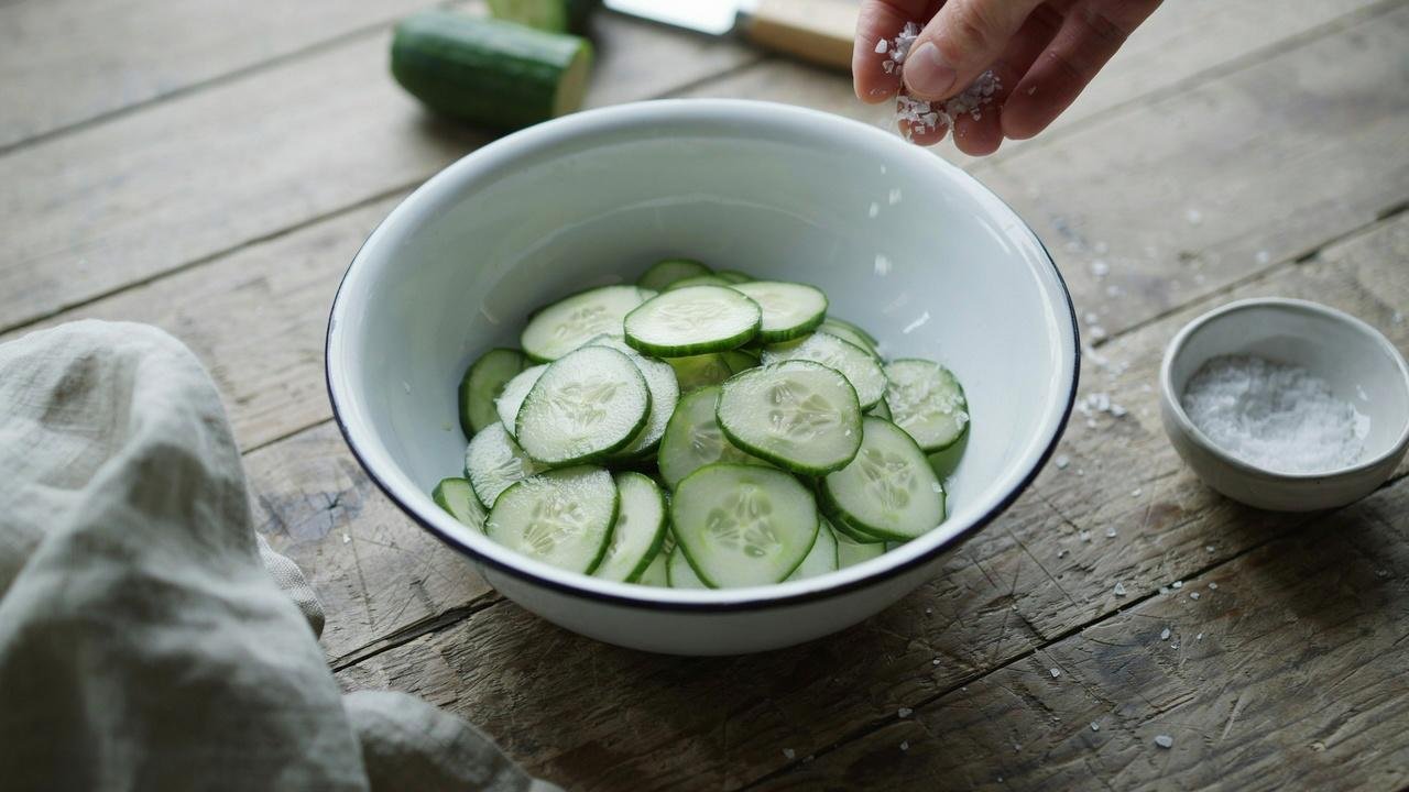 Sliced cucumbers in a white colander with salt being sprinkled over them by hand.