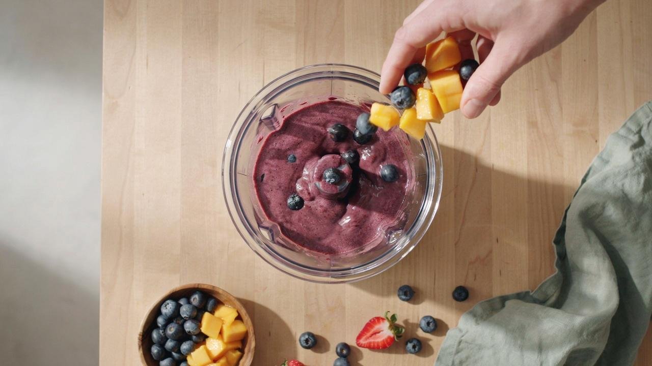 Frozen berries and mango pieces added to thick purple vegan acai bowl mixture in blender.