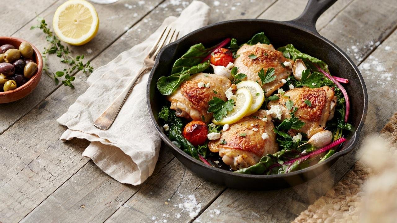 Golden Mediterranean chicken with sautéed spinach, kale, and Swiss chard in a rustic cast-iron skillet, garnished with lemon, parsley, and feta cheese on a wooden table.