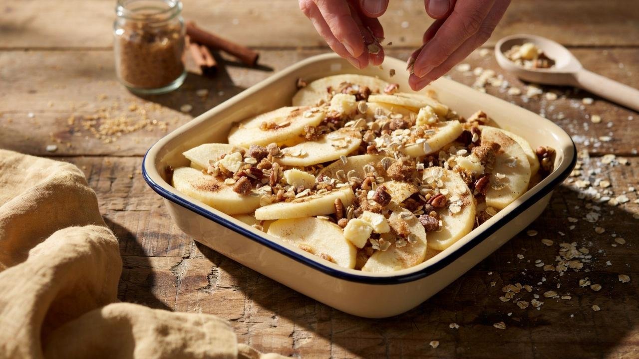 Overhead shot of hands gently spreading golden oat crumble mixture over a layer of sliced cinnamon apples in a baking dish.