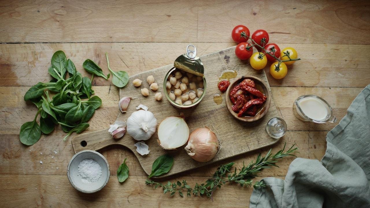 Fresh ingredients for creamy Tuscan chickpeas including chickpeas, spinach, tomatoes, garlic, herbs, and cream on a kitchen counter.