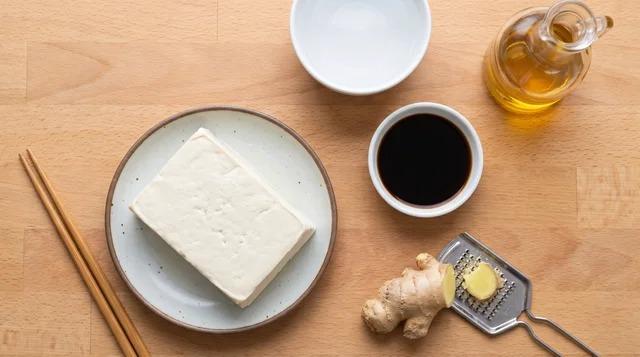 Fresh tofu block beside cool water, soy sauce, ginger, and sesame oil